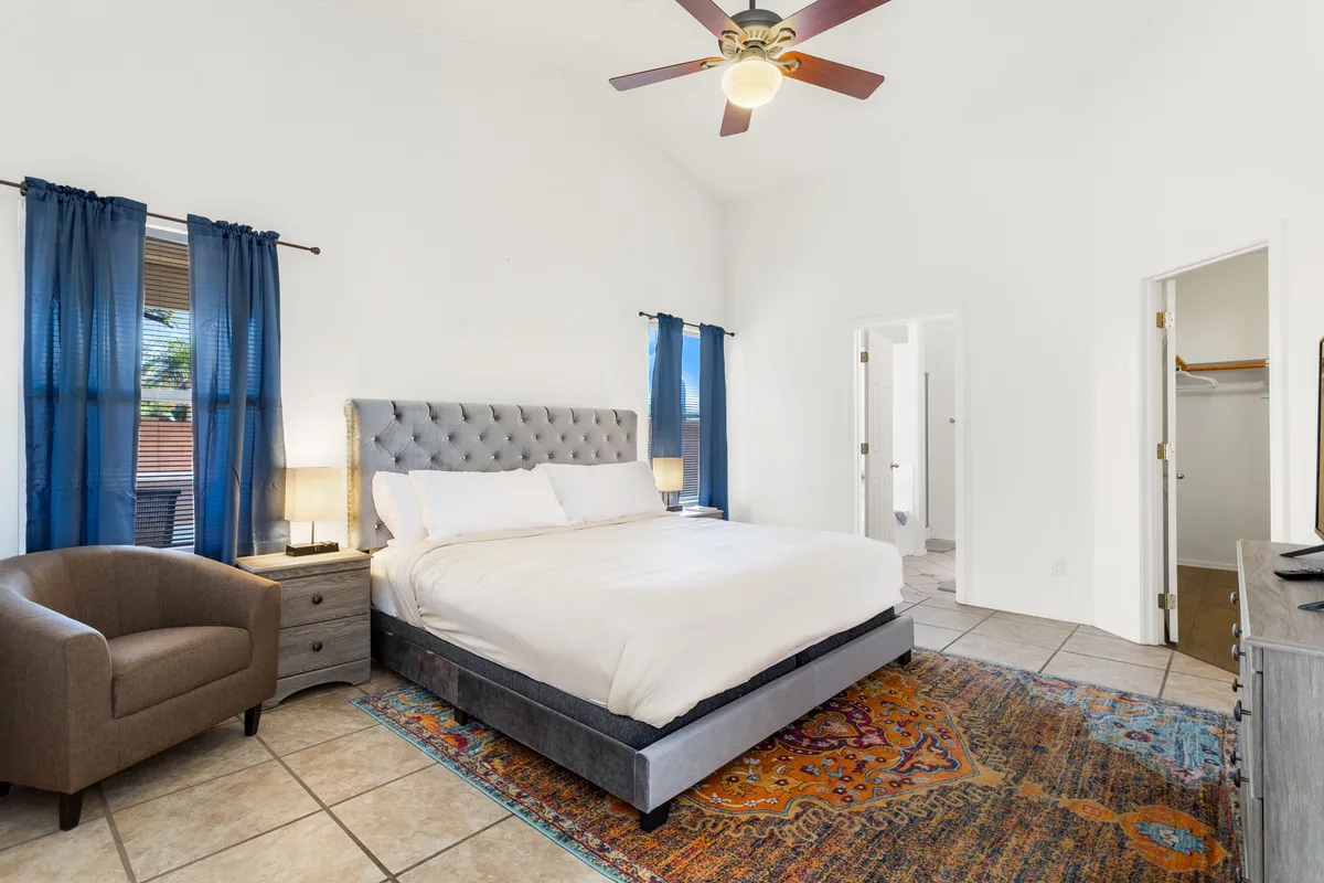 This image features a serene bedroom with a large, comfortable bed centered on a soft area rug. Natural light pours in through windows, illuminating a cozy chair and nightstands on either side, creating an atmosphere ideal for relaxation and quiet moments. The decor is calm and inviting, reflecting a restful retreat.