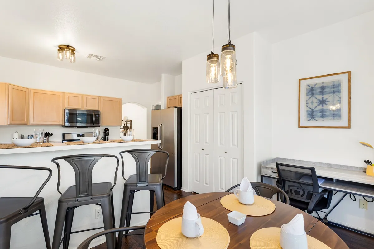 A bright, open area featuring a kitchen island, dining table, pendant lighting, and artwork, with large windows letting in natural light—the room feels inviting and open to the outside.