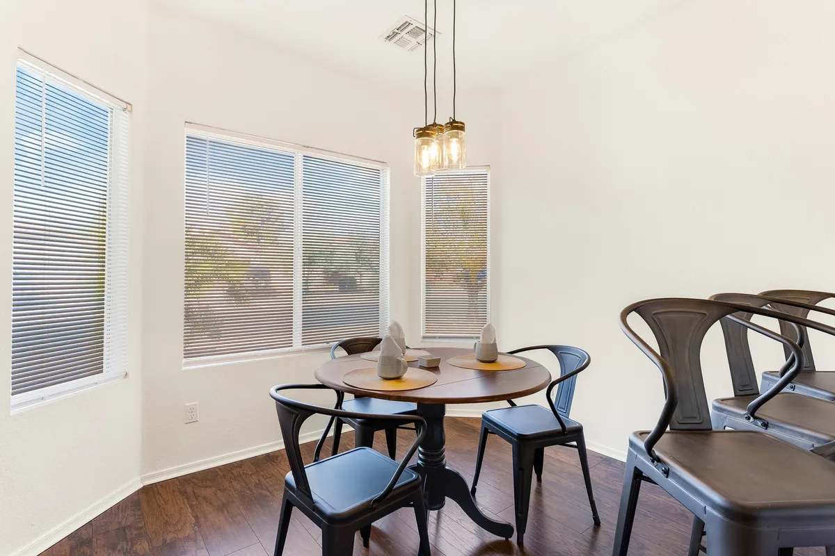 A cozy, sunlit dining area featuring a round table surrounded by comfortable chairs. The room is enhanced by hanging light fixtures and large windows with blinds, creating a warm environment ideal for gathering with family or friends.