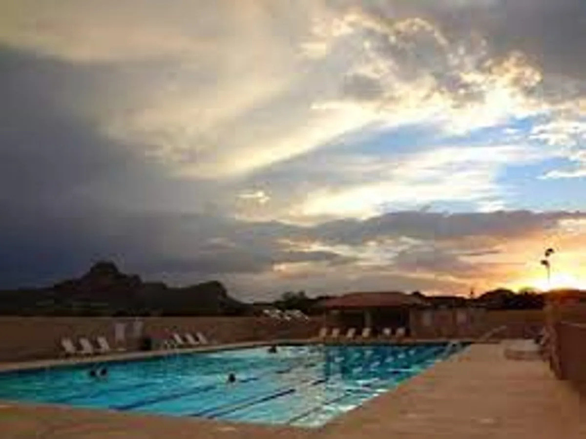 A tranquil community swimming pool set against a backdrop of mountains and a colorful sunset sky. The inviting water reflects the warm hues of evening, offering a resort-like atmosphere ideal for relaxation and socializing.
