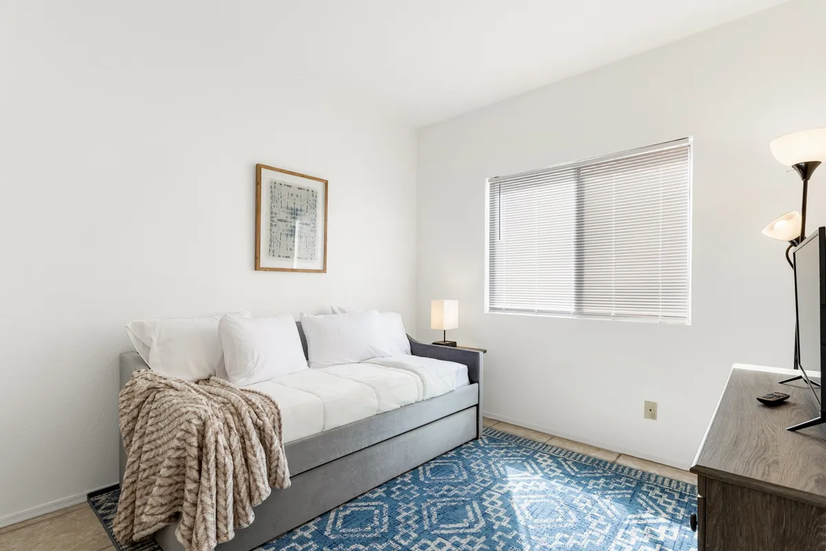 A cozy nook with a daybed dressed in soft textiles, accented by wall art and a warm lamp. The rug underfoot and the inviting setup make it perfect for relaxing, reading, or napping, echoing a welcoming retreat vibe. The space feels private and snug—ideal for children or solo travelers.