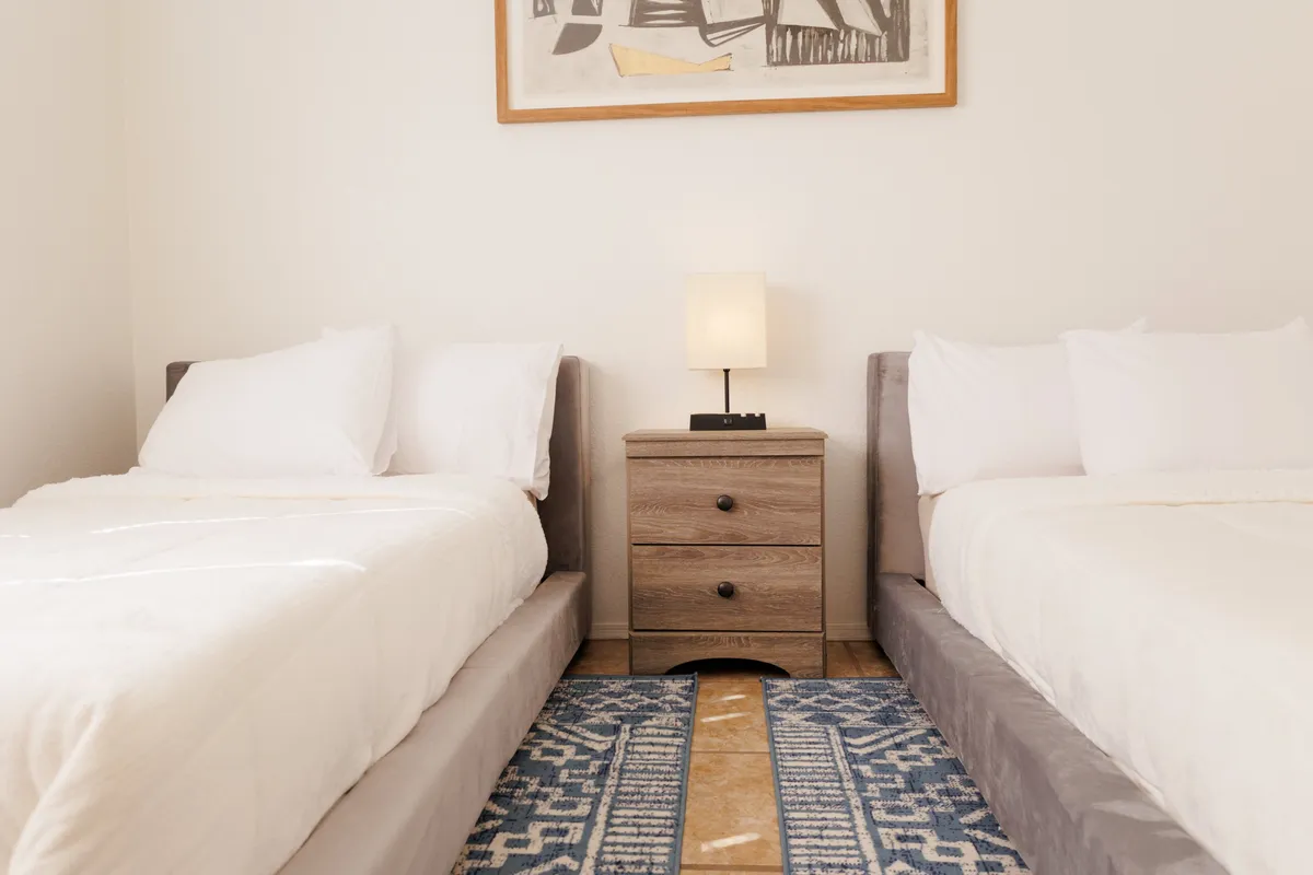 This photo showcases a flexible bedroom setup with two twin beds, a nightstand with a lamp between them, and uplifting wall art. The setting feels inviting for both children and adults, emphasizing adaptability for various guest needs. The room exudes comfort and a sense of home away from home.